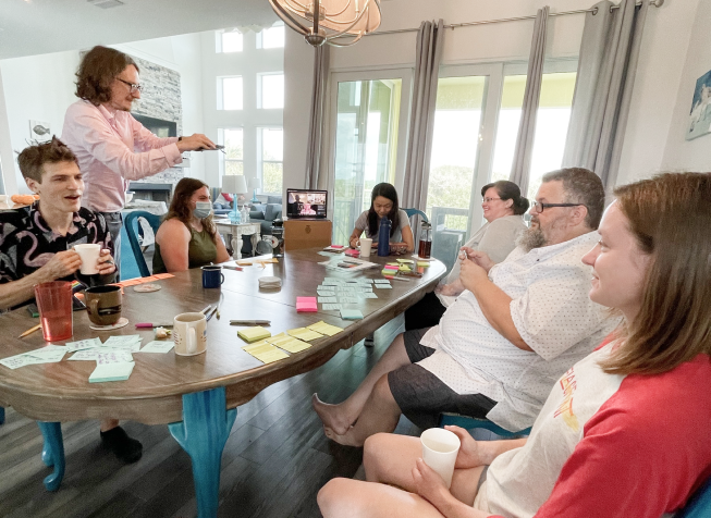 A group of Flagrant team members, including one person joining via laptop are gathered around a large table in conversation. The table is covered in sticky notes, coffee mugs, and pencils, as well as other tools to enable collaboration.