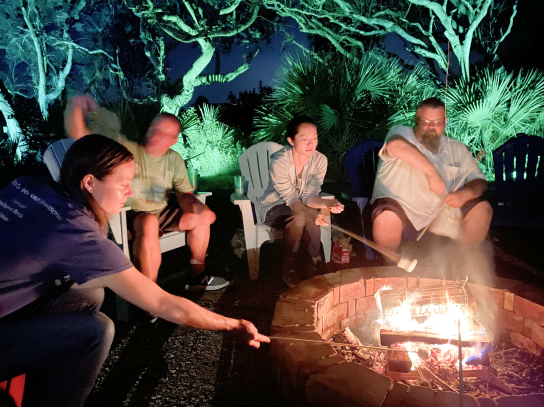 A group of Flagrant team members are gathered around a firepit, roasting smores.