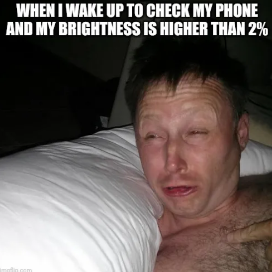 A man in a dark room with a bright light shining on his face lifts his head off a pillow with raised eyebrows and very squited eyes. Above him in meme-style text is written "When I wake up to check my phone and my brightness is higher than 2%."