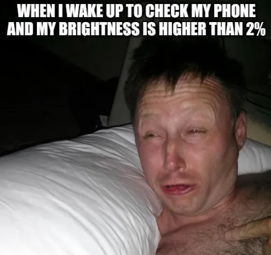 A man in a dark room with a bright light shining on his face lifts his head off a pillow with raised eyebrows and very squited eyes. Above him in meme-style text is written "When I wake up to check my phone and my brightness is higher than 2%."