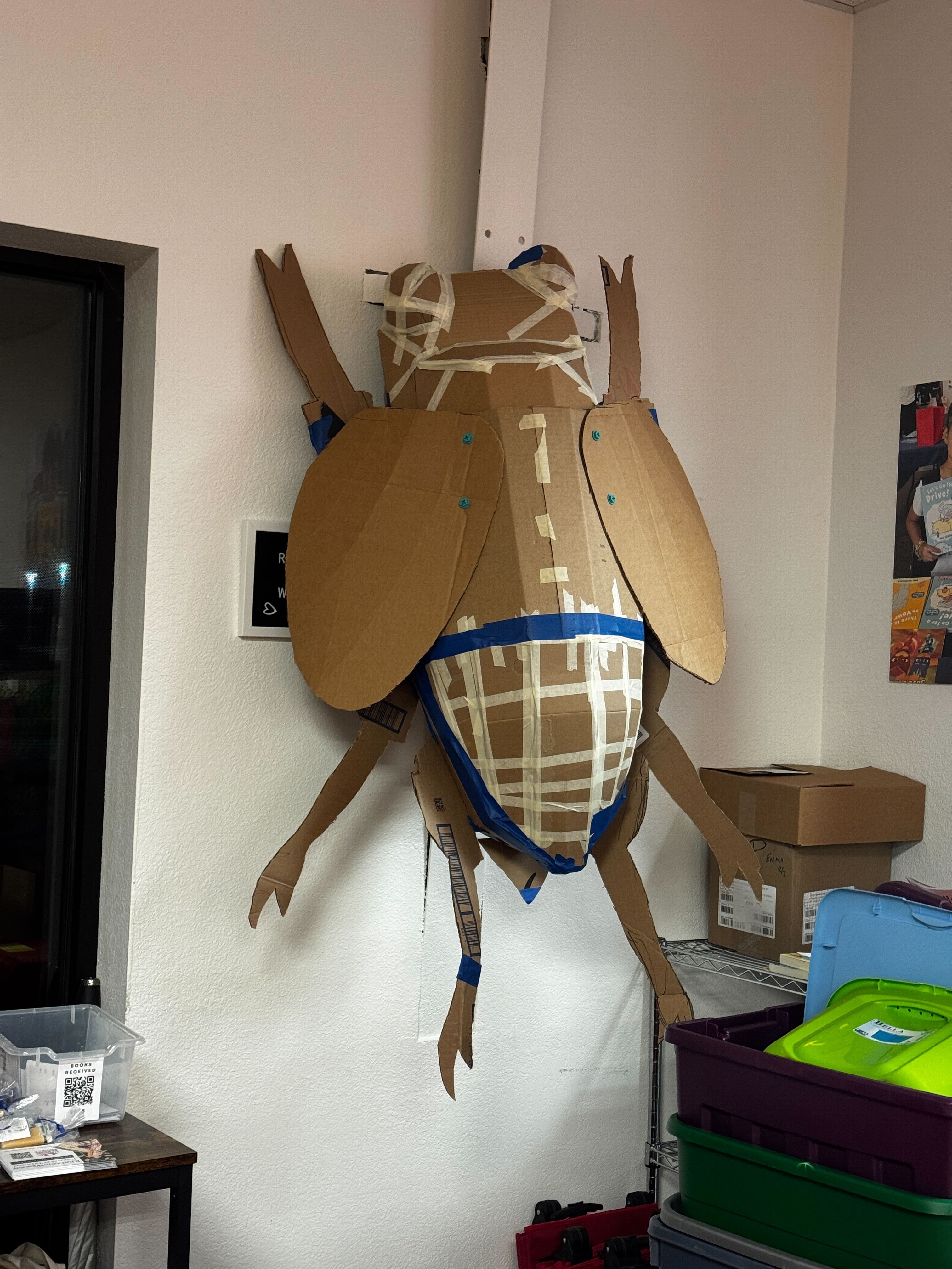 Large bug made out of taped together cardboard hanging on the wall.